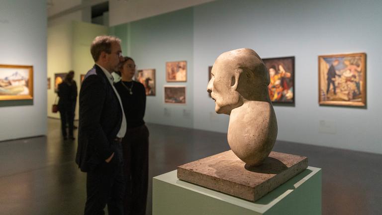 In einem Ausstellungsraum betrachten Menschen an der Wand hängende bunte Gemälde. Im Vordergrund steht die Büste eines Mannes.