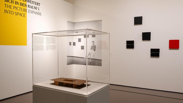 In einem Ausstellungsraum steht ein Glaskasten mit einem Exponat. An der Wand hängen Bilder und man sieht eine Schrift an der Wand.