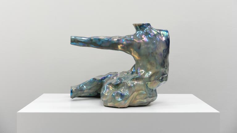 In einem Ausstellungsraum sieht man eine abstrakte, schillernde Figur, die an einen Menschen erinnert.