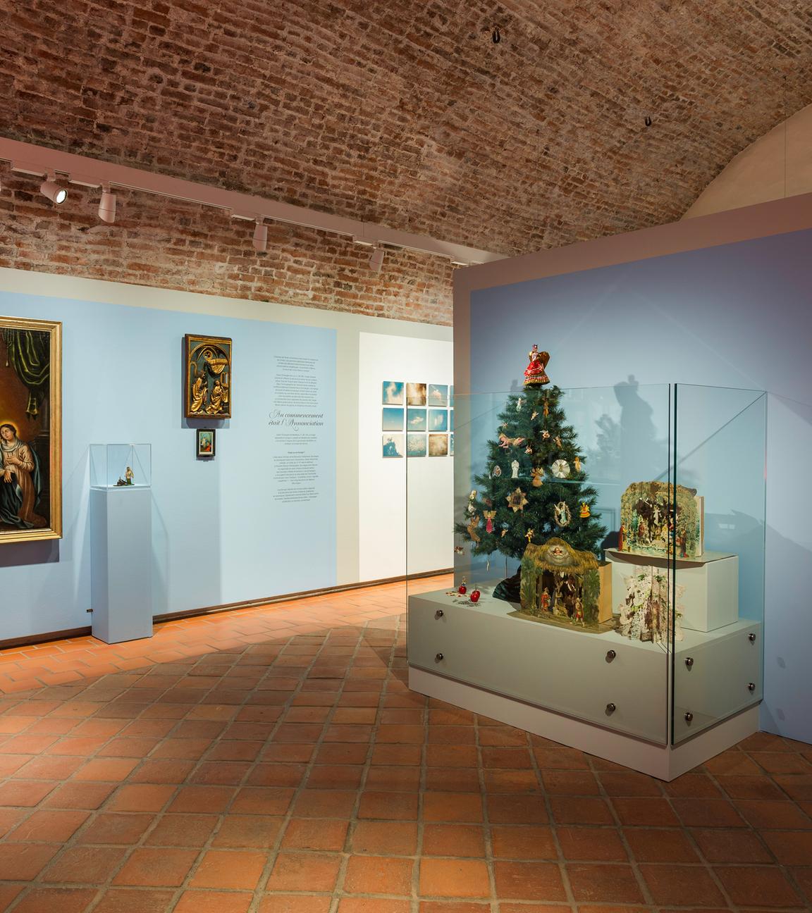 Zu sehen ist ein Raum der Ausstellung. Links an der Wand hängen mehrere Bilder, auf denen Engel zu erkennen sind. Rechts steht ein kleiner geschmückter Weihachtsbaum in einer Vitrine vor einer Wand.