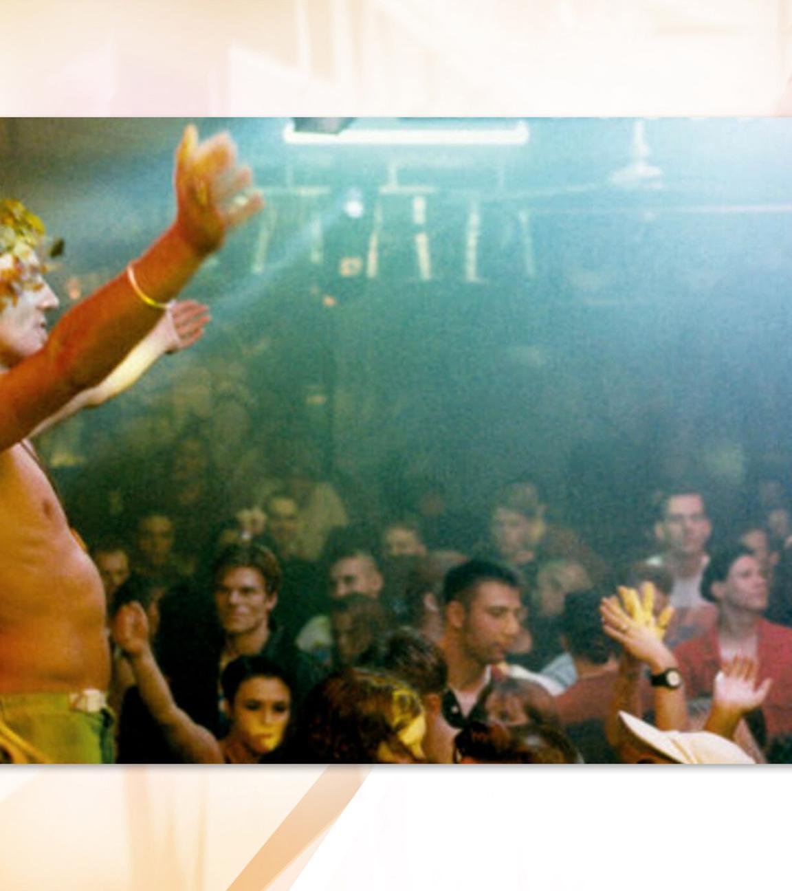 Ein Mann der einen Blumenkranz auf dem Kopf trägt, steht in einer Menge tanzender Menschen in einer Diskothek und hebt die Arme empor. 