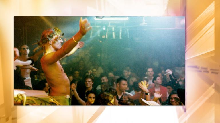 Ein Mann der einen Blumenkranz auf dem Kopf trägt, steht in einer Menge tanzender Menschen in einer Diskothek und hebt die Arme empor. 