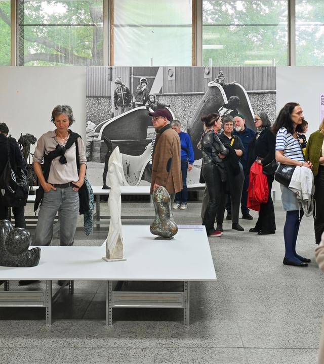 In einem Ausstellungsraum schauen Menschen sich die Plastiken, die auf Tischen stehen und großformatige Schwarz-weiß-Fotografien an den Wänden an.