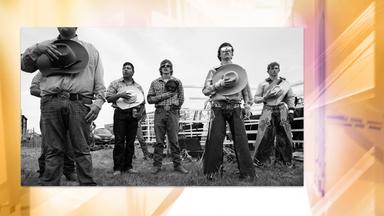 Auf einer Schwarz-Weiß-Fotografie sieht man mehrere breitbeinig nebeneinander stehende Cowboys , die sich jeweils ihren Hut auf die Brust halten.
