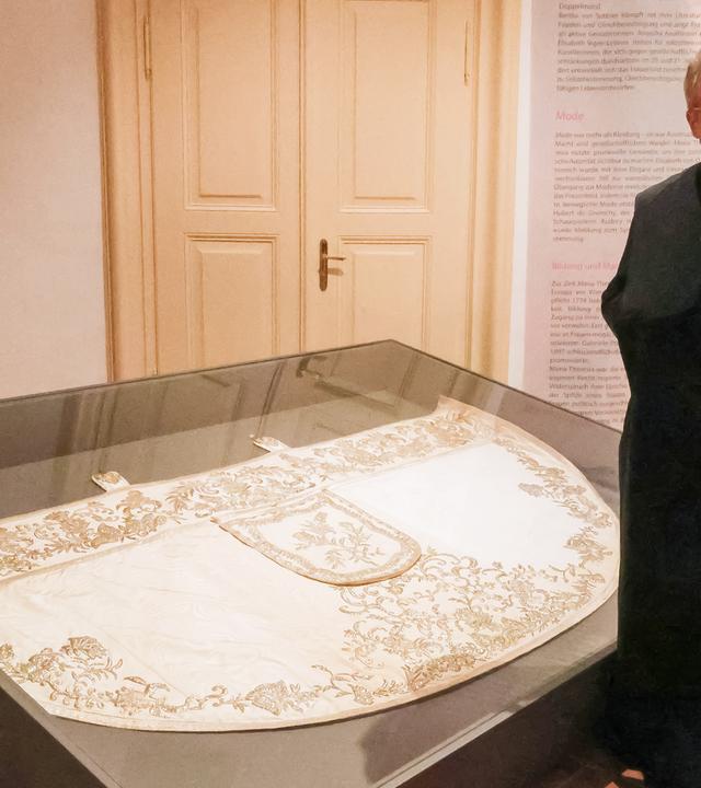 Ein Mann steht neben einer Glasvirtrine, in der ein historischer weißer Umhang mit Goldstickerei liegt.