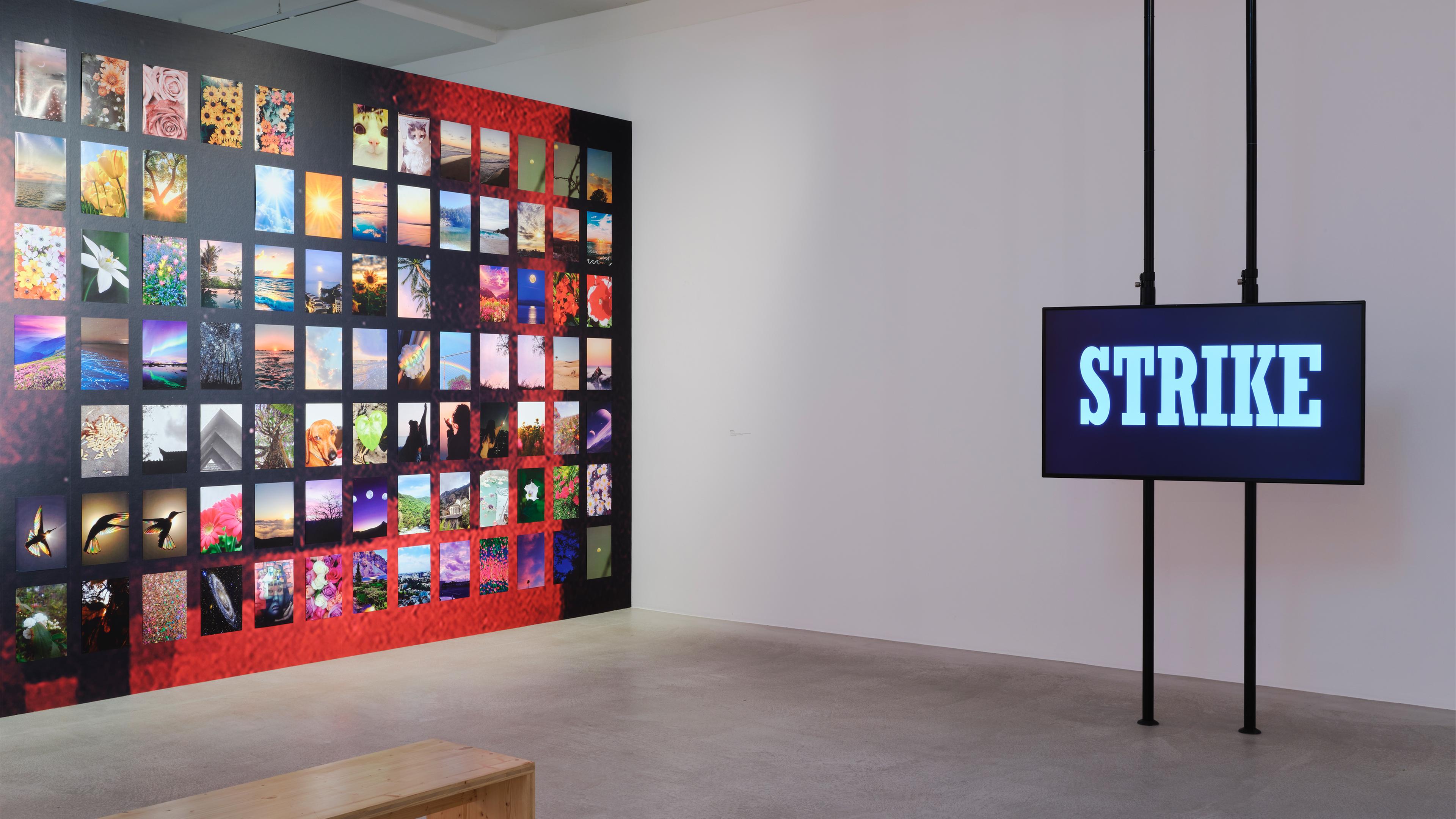 In einem Ausstellungsraum befinden sich links viele, bunte Fotos an einer Wand. Rechts ist ein Schild mit dem Wort "Strike" zu sehen.