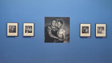 In einem Ausstellungsraum sieht man Schwarz-Weiß-Fotografien an einer blauen Wand. Die Fotografien zeigen tätowierte Menschen.