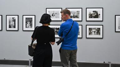 In einem Ausstellungsraum schauen eine Frau und ein Mann sich Schwarz-Weiß-Fotografien an einer Wand an.