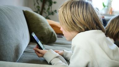 21.04.2025, Bayern, Roth: ILLUSTRATION - Ein Junge liegt auf einem Sofa und guckt auf sein Smartphone. (gestellte Szene)