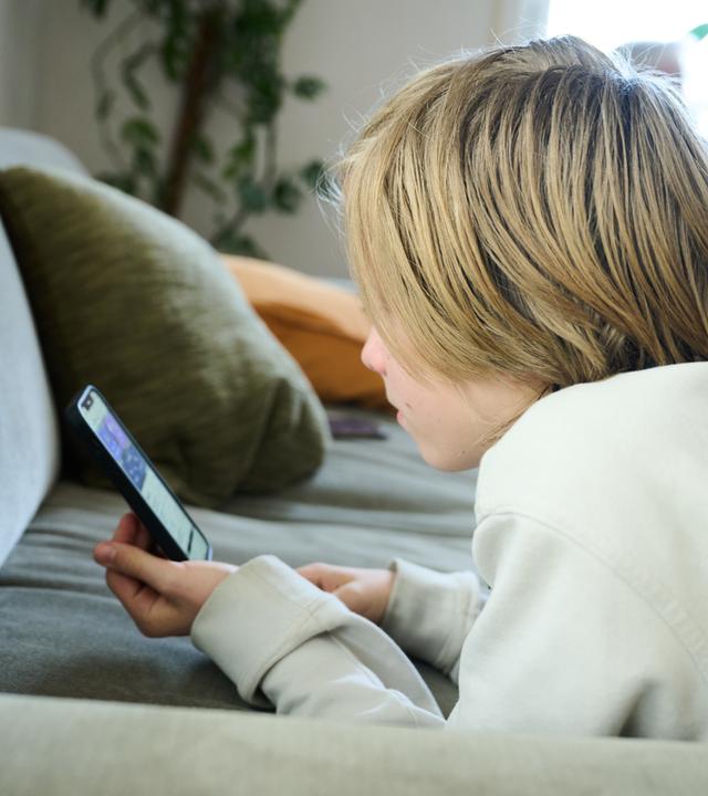 21.04.2025, Bayern, Roth: ILLUSTRATION - Ein Junge liegt auf einem Sofa und guckt auf sein Smartphone. (gestellte Szene)