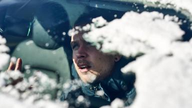 Ein Mann sitzt im Auto und blickt durch die verschneite Windschutzscheibe.