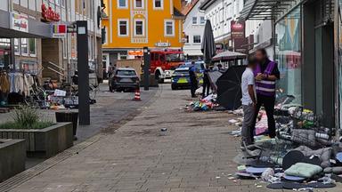 Das Bild zeigt die Fußgängerzone in Hofgeismar, die durch den Autofahrer verwüstet wurde. An der Seite stehen Menschen, hinten sieht man das Fahrzeug des Rasers und ein Polizeiauto mit Polizisten.
