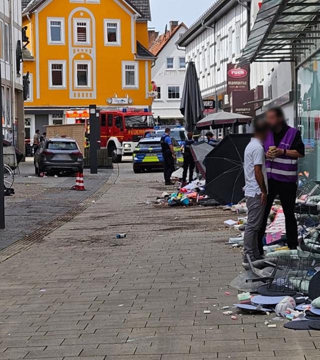 Das Bild zeigt die Fußgängerzone in Hofgeismar, die durch den Autofahrer verwüstet wurde. An der Seite stehen Menschen, hinten sieht man das Fahrzeug des Rasers und ein Polizeiauto mit Polizisten.