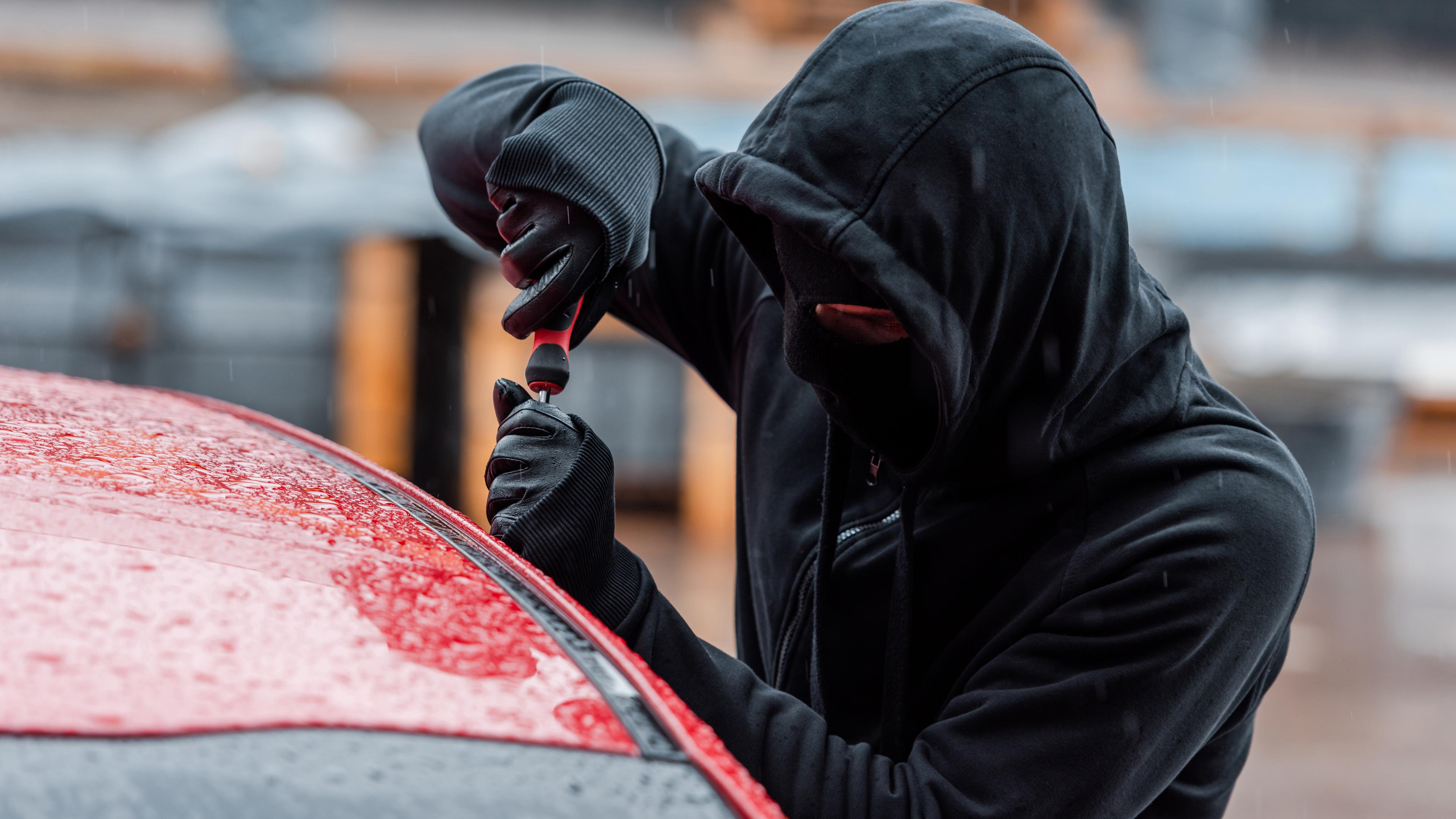 Ein Mann in schwarzer Kleidung mit Handschuhen und Maske versucht eine Autotür mit einem Schraubenschlüssel zu öffnen.