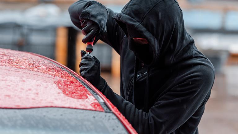 Ein Mann in schwarzer Kleidung mit Handschuhen und Maske versucht eine Autotür mit einem Schraubenschlüssel zu öffnen.