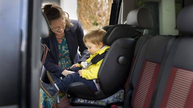 Eine Mutter schnallt ihr Kind, das in einem Autokindersitz sitzt, an.