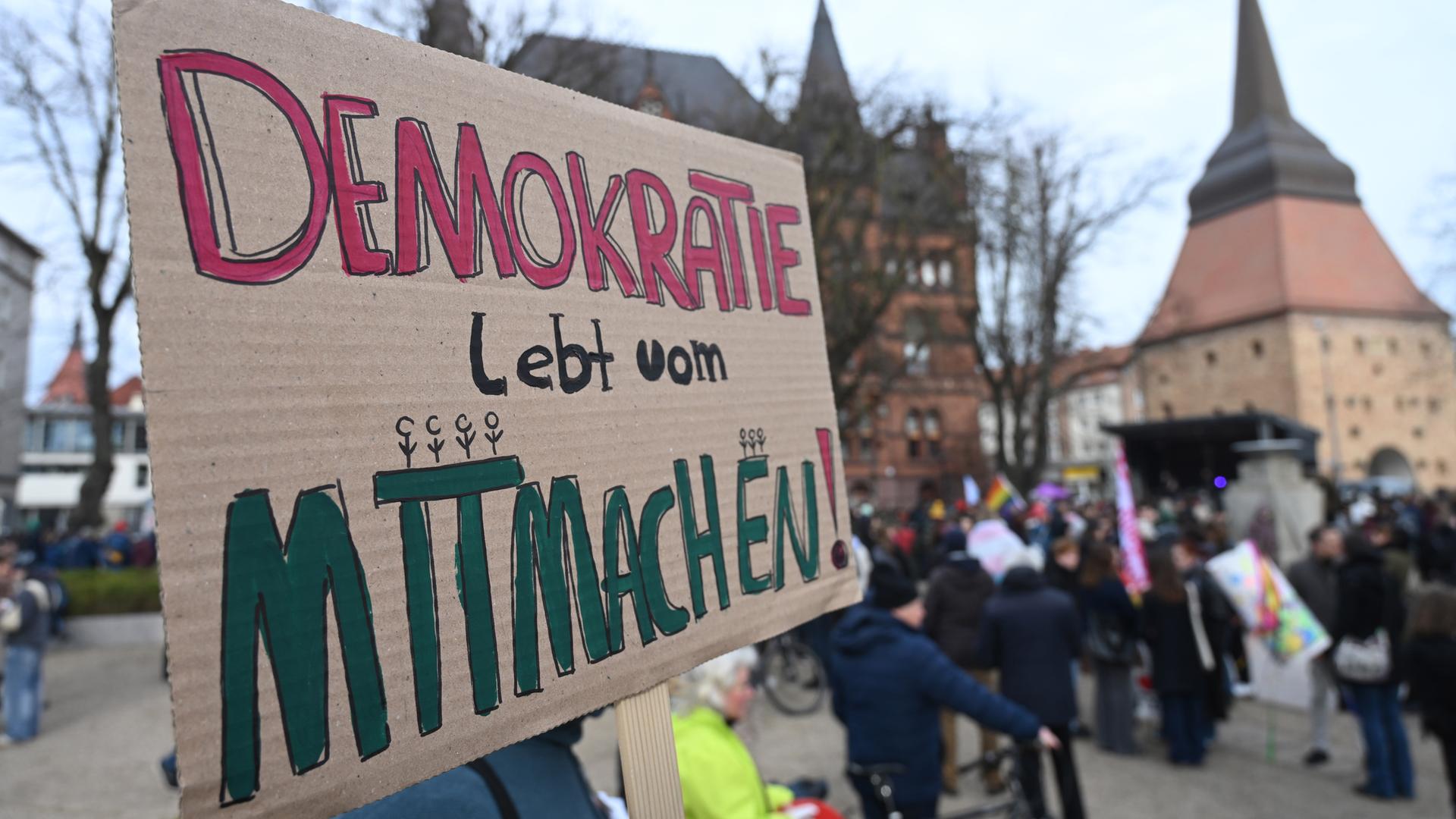 22.02.2025, Mecklenburg-Vorpommern, Rostock: Teilnehmer einer Demonstration und Kundgebung unter dem Motto "Solidarität statt Ausgrenzung" zeigen in der Innenstadt ein Plakat mit der Aufschrift "Demokratie lebt vom Mitmachen!".
