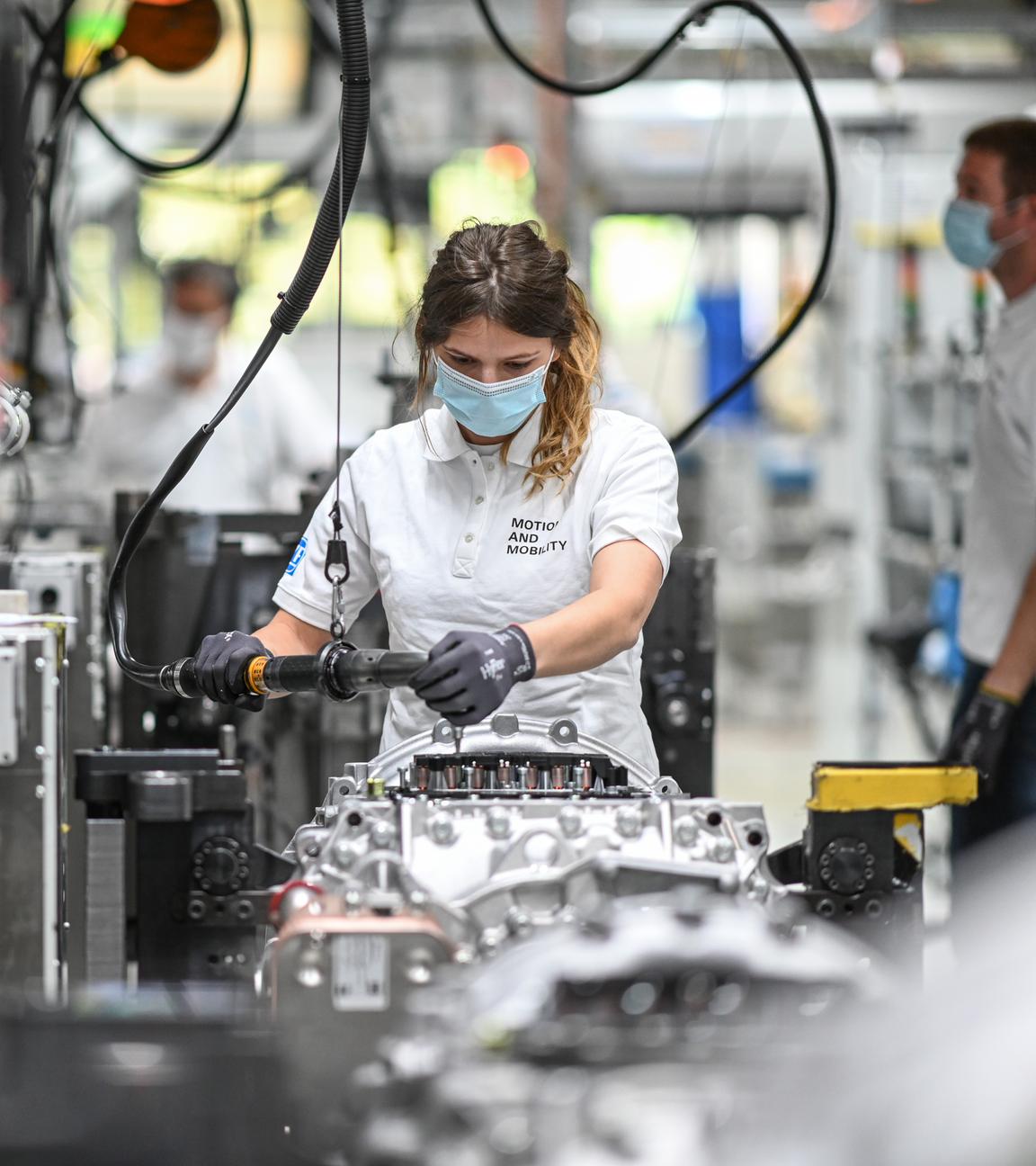 Eine Person in weißem Oberteil und mit Maske arbeitet an silbernen Industrieteilen.