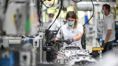 Eine Person in weißem Oberteil und mit Maske arbeitet an silbernen Industrieteilen.