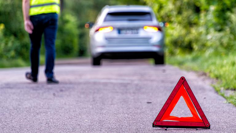 Zu sehen ist ein auf einer Landstraße stehendes Auto, ein Mensch in einer Warnweste, der dem Betrachter den Rücken zukehrt, sowie im Vordergrund ein Warndreieck
