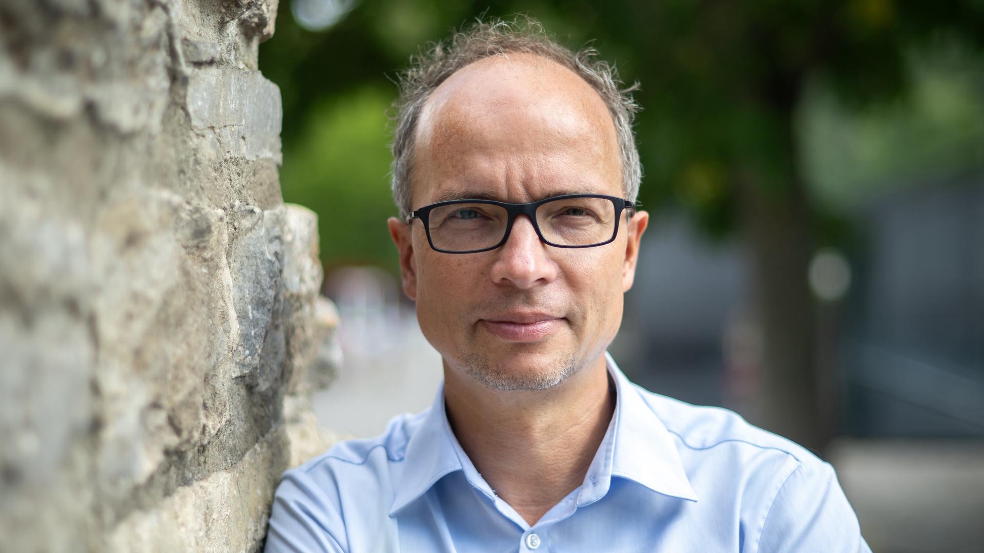 Martin Debes trägt ein hellblaues Hemd und schaut, an eine Steinwand gelehnt, direkt in die Kamera.