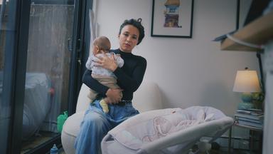 Eine Frau sitzt und hält ein Baby auf dem Arm, vor ihr steht eine Babywippe