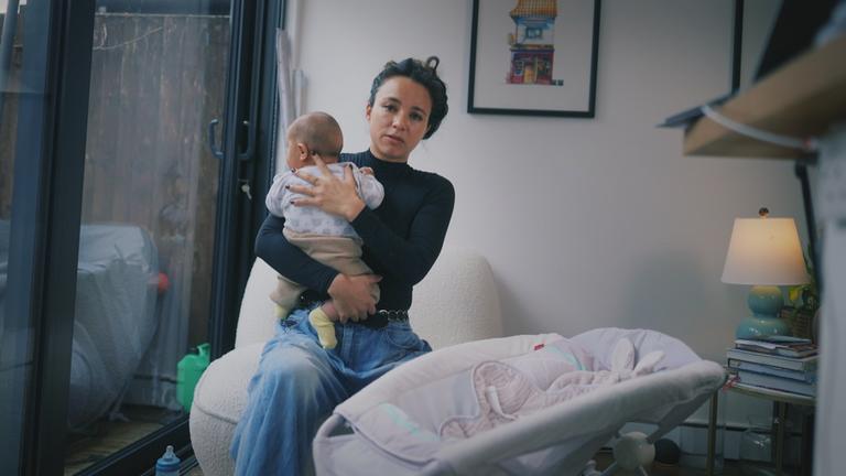 Eine Frau sitzt und hält ein Baby auf dem Arm, vor ihr steht eine Babywippe