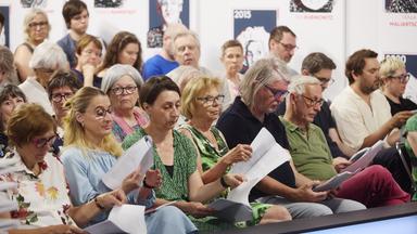 Das Bild zeigt eine Veranstaltung im Rahmen der 50. Tage der deutschsprachigen Literatur, speziell den Ingeborg-Bachmann-Preis 2026. Im Vordergrund sitzen mehrere Personen in Reihen auf Stühlen. Die Zuhörer halten Papiere in den Händen.