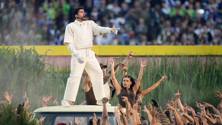 Bad Bunny bei seiner Halbzeitshow im weißen Anzug, steht auf dem Dach eines Autos und singt.