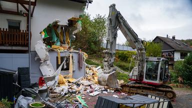 Links steht ein Wohnhaus mit einer großflächig aufgerissenen Außenwand. Die Öffnung reicht über zwei Geschosse und gibt den Blick auf Innenräume, gelbes Dämmmaterial und herabhängende Folien frei. Rechts daneben steht ein grau-roter Bagger.