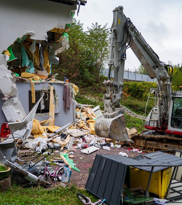 Links steht ein Wohnhaus mit einer großflächig aufgerissenen Außenwand. Die Öffnung reicht über zwei Geschosse und gibt den Blick auf Innenräume, gelbes Dämmmaterial und herabhängende Folien frei. Rechts daneben steht ein grau-roter Bagger.