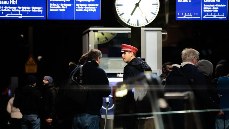 Menschen stehen an mit einem DB-Mitarbeiter an einem Bahnhof.