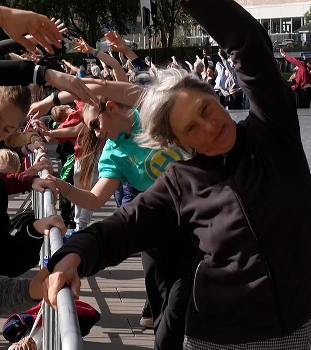Menschen machen an einer Absperrung Ballettübungen. Im Vordergrund dehnen sich zwei Frauen seitlich mit erhobenem Arm. 