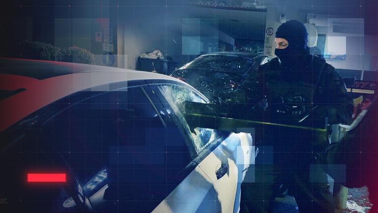 Zwei Vermummte Polizisten stehen neben einem geparktem Pkw, in dem sich mutmaßlich eine Waffe befindet. Einer von ihnen öffnet die Seitenscheibe des Fahrzeuges gewaltsam mit einer Ramme