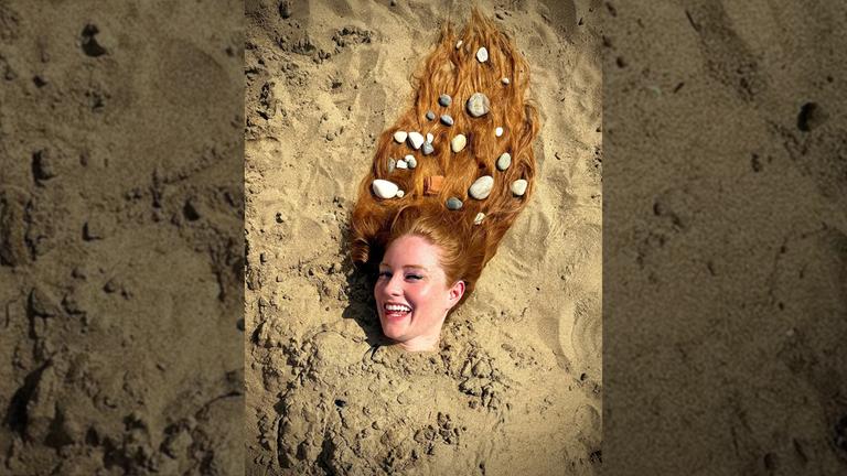 Zu sehen ist, wie Barbara Meier am Strand eingebuddelt ist. Ihr Kopf ragt hervor und schaut in die Kamera.