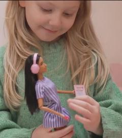 Ein Mädchen hat eine Barbie-Puppe in der Hand und lächelt. Neben ihr kniet eine Frau, die das Mädchen anschaut.