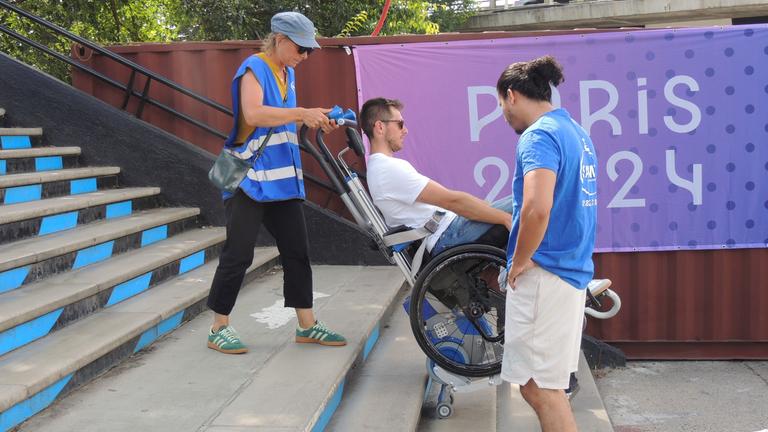 Ein Mann im Rollstuhl wird mit Hilfe von zwei Helfern ein Treppe hochgehoben.