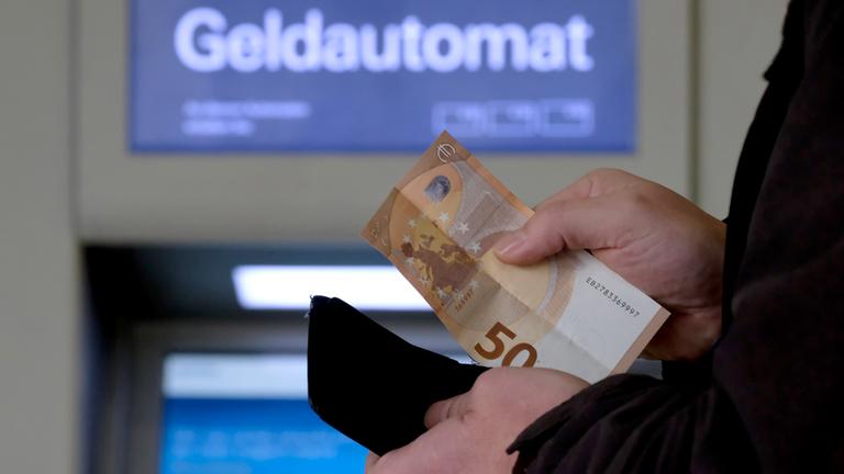 Eine Geldbörse mit einem 50-Euro-Schein wird vor einen Geldautomaten gehalten.