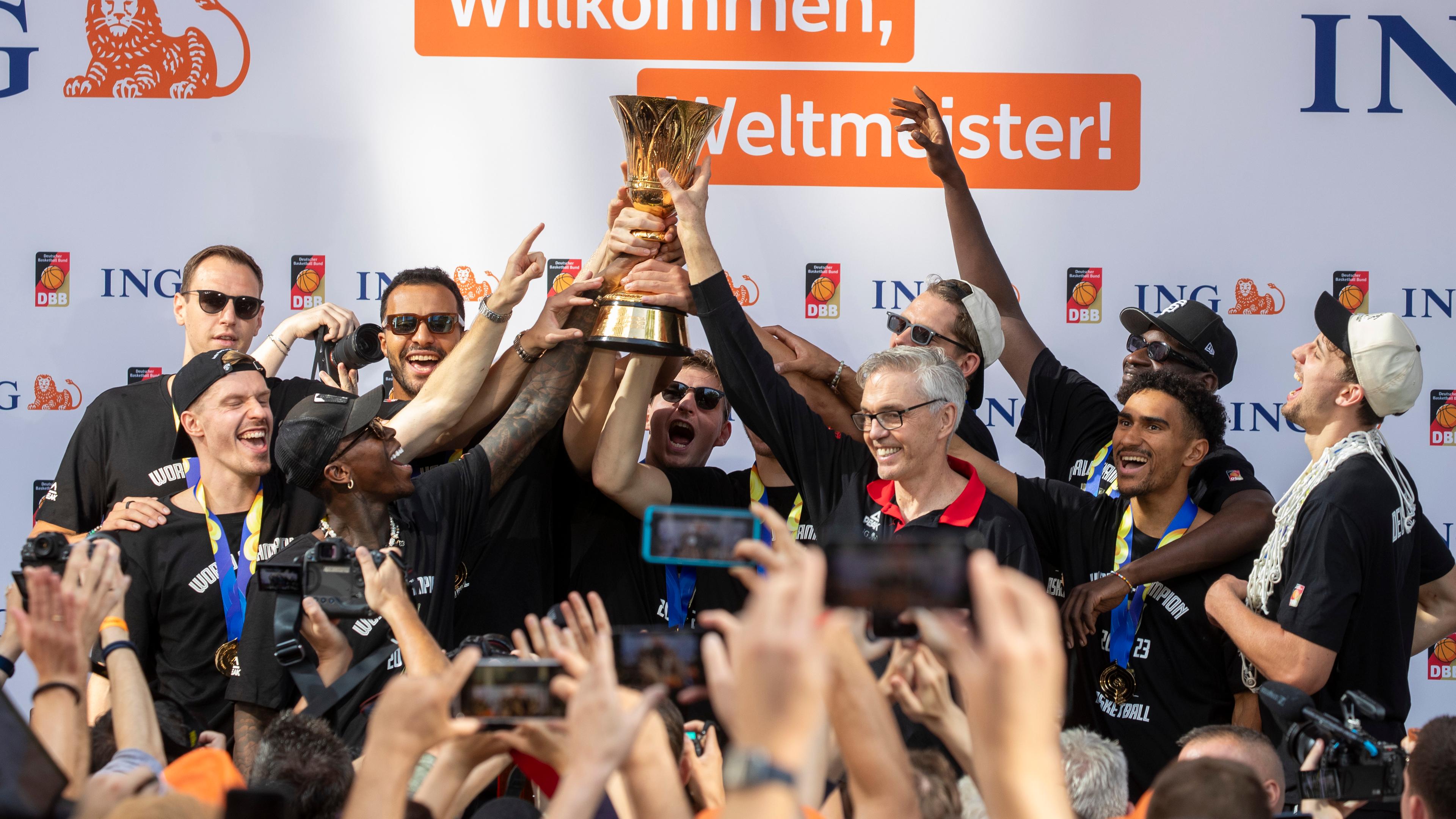 12.09.2023, Hessen, Frankfurt/Main: Dennis Schröder (3.v.l) und Trainer Gordon Herbert (4.v.r) Halten den Pokal in die Luft und sind umringt von der Mannschaft. 