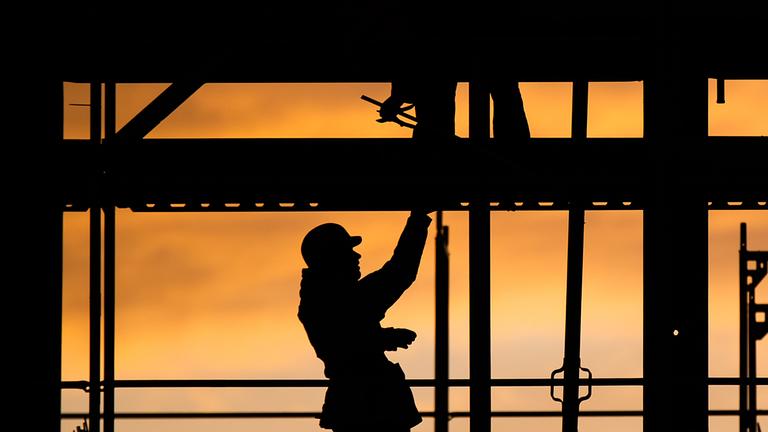 Die Silhouette eines Bauarbeiters zeichnet sich auf einer Baustelle vor dem verfärbten Morgenhimmel ab. 