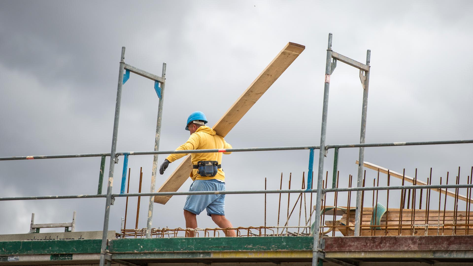 Ein Bauarbeiter trägt auf einer Baustelle auf einem Hausdach ein Brett.