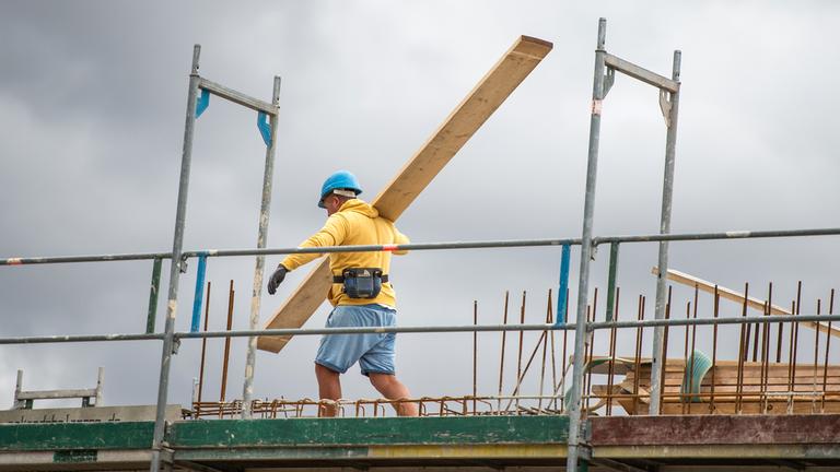 Ein Bauarbeiter trägt auf einer Baustelle auf einem Hausdach ein Brett