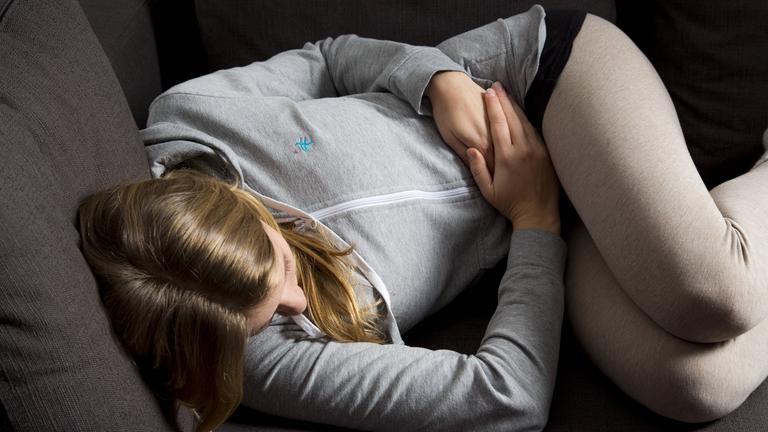 Frau liegt auf dem Sofa und hält sich den Bauch.