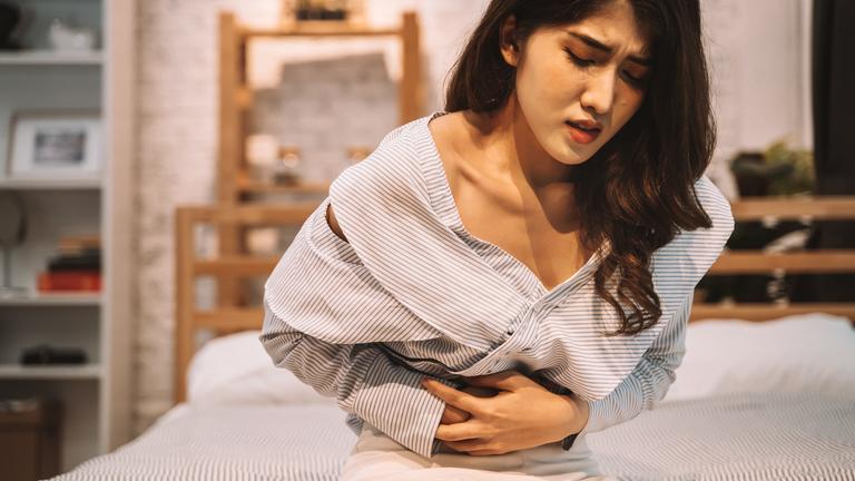 Eine junge Frau sitzt auf einem Bett und hält sich vor Schmerzen den Bauch.