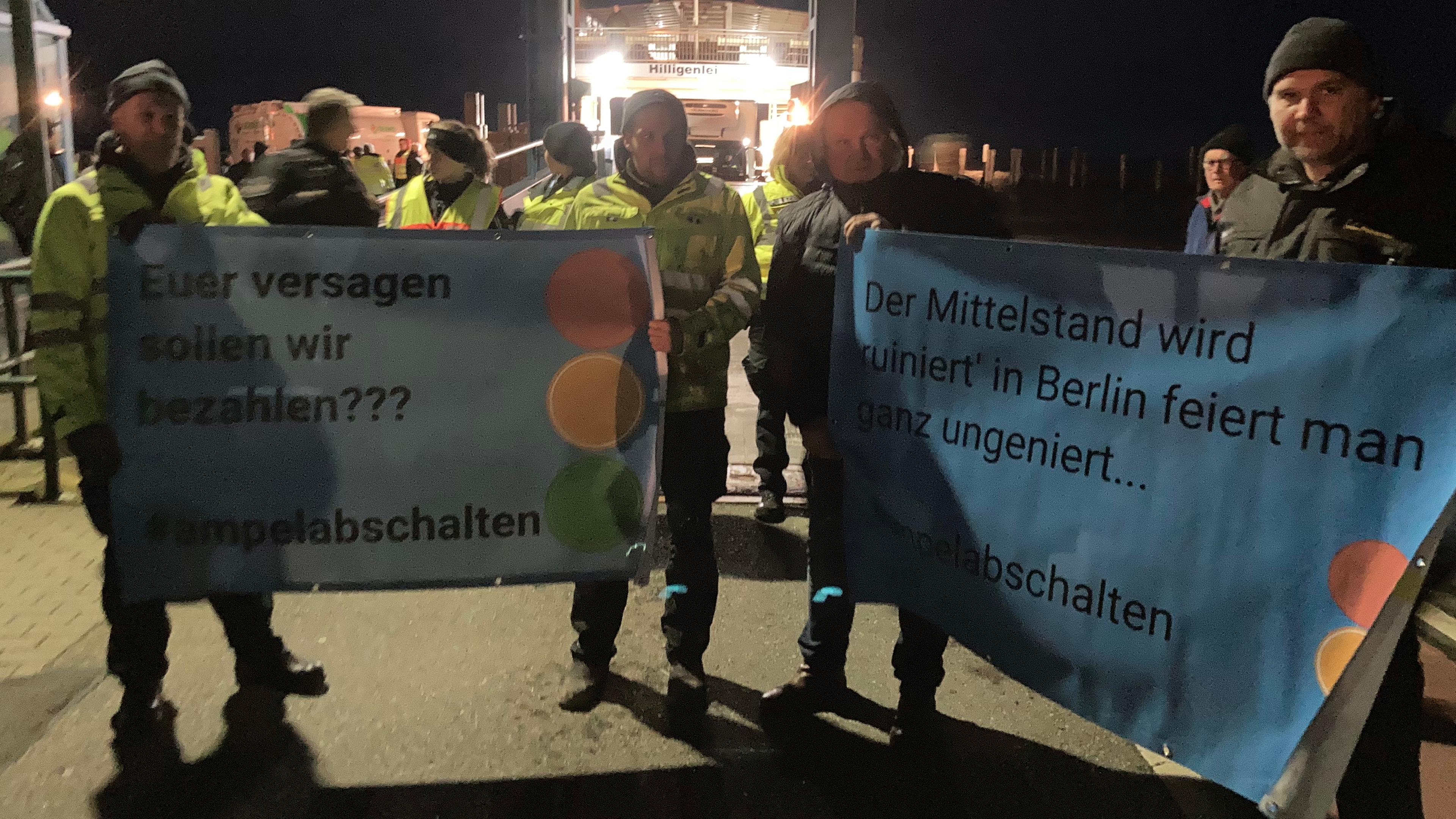  Landwirte stehen mit Plakaten mit der Aufschrift "Euer Versagen sollen wir bezahlen??? #ampelabschalten" am Fähranleger. 