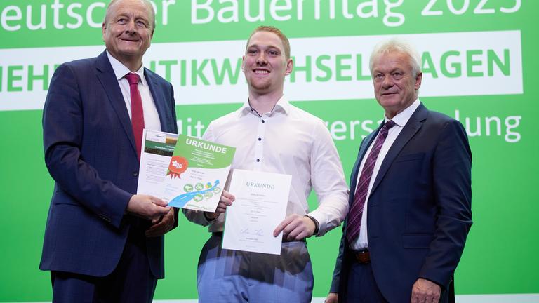 oachim Rukwied (l), Präsident des Deutschen Bauernverbands , und Alois Rainer (CSU), Bundeslandwirtschaftsminister, gratulieren Felix Winkler aus Rheinland Pfalz zum Sieg beim Berufswettbewerb der Deutschen Landjugend in der Sparte Weinbau auf einer Bühne.