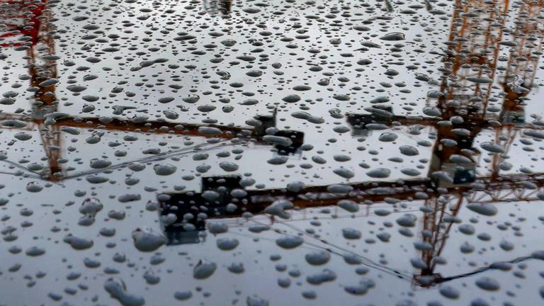 Baukräne auf dem Gelände eines Kranservice spiegeln sich auf einem mit Regentropfen bedeckten Auto. 