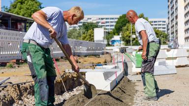 Zwei Bauarbeiter sind auf einer Baustelle, der Himmel ist blau.