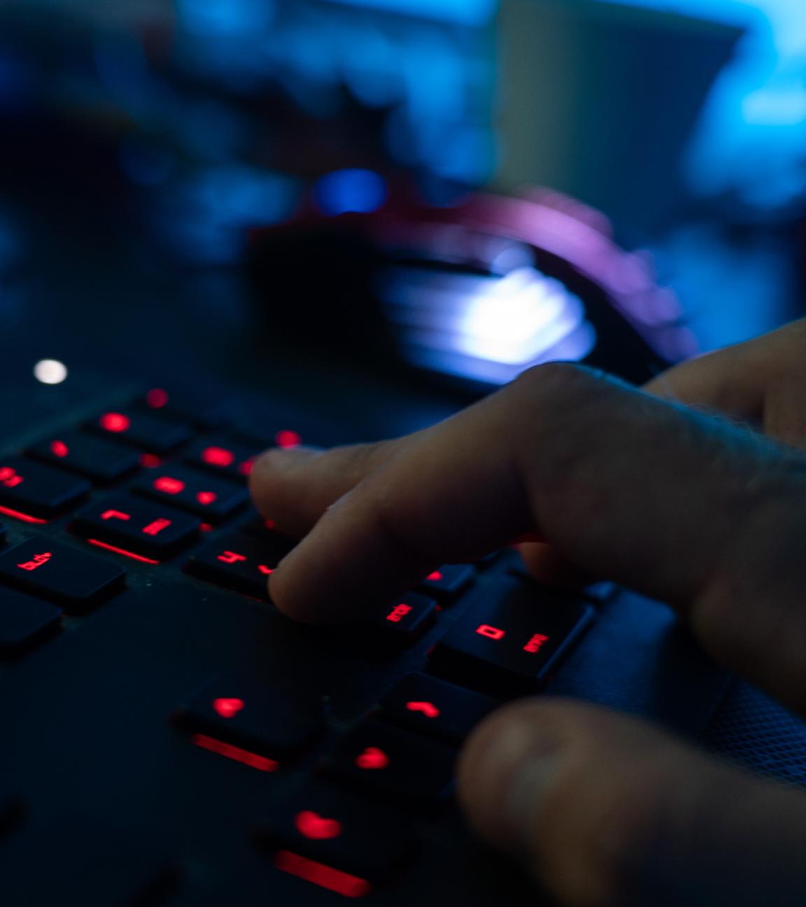 Eine rot beleuchtete Tastatur in dunklem, bläulichen Licht, eine rechte Hand schreibt auf der Tastatur.