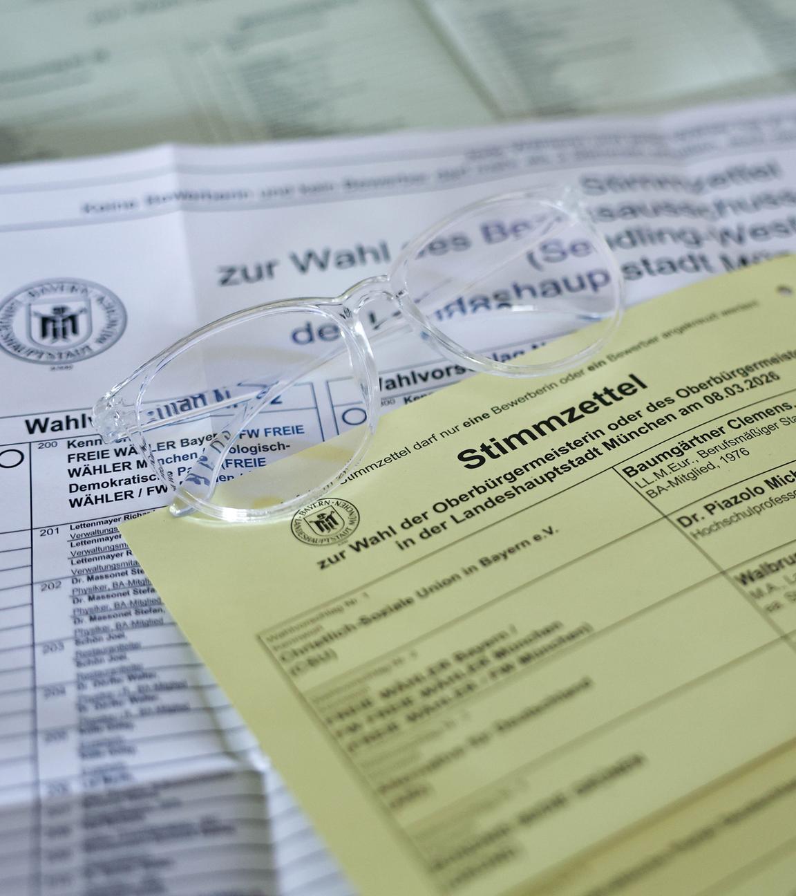 Briefwahlunterlagen für die Kommunalwahl in Bayern 2026 liegen auf dem Tisch. Darauf liegt eine Brille mit durchsichtigem Rahmen.